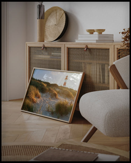 Ein gerahmtes Poster von einem Leuchtturm auf Sylt, der hinter grasbedeckten Dünen hervorschaut, an einer Holzkommode angelehnt und stehend auf dem Holzboden