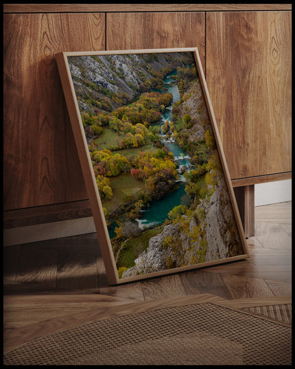 Ein gerahmtes Poster von einem Fluss der sich, umgeben von mehreren Büschen, durch eine Berglandschaft erstreckt, an einer Holzkommode angelehnt und stehend auf dem Holzboden