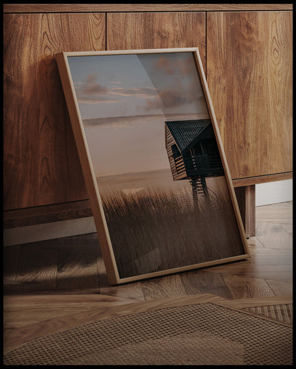 Ein gerahmtes Poster von einer Hütte am Strand in den Dünen bei Sonnenuntergang, an einer Holzkommode angelehnt und stehend auf dem Holzboden