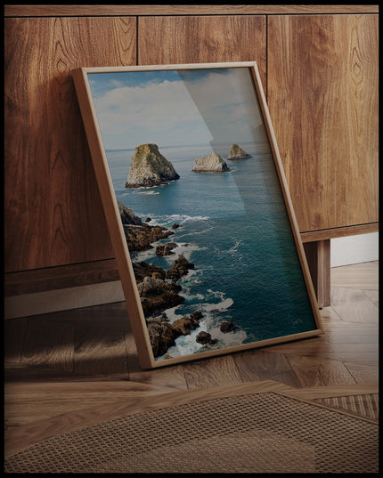 Ein gerahmtes Poster vom Blick auf das blaue Meer und mehrere Felsformationen die aus dem Wasser blicken, an einer Holzkommode angelehnt und stehend auf dem Holzboden