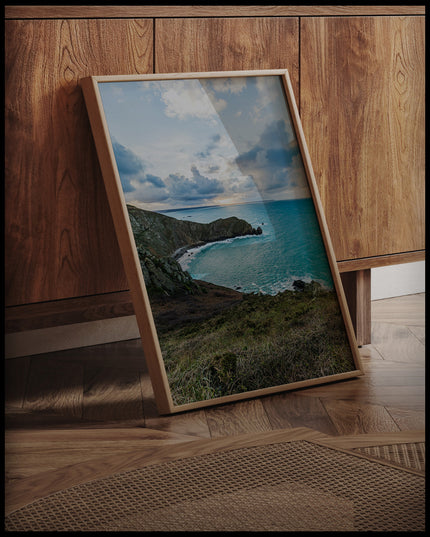 Ein gerahmtes Poster vom Blick einer Bucht über das blaue Meer, an einer Holzkommode angelehnt und stehend auf dem Holzboden