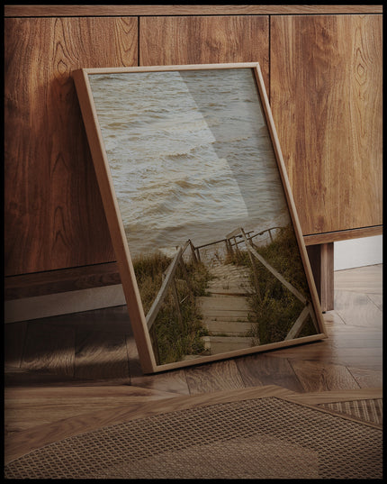 Ein gerahmtes Poster von einer Holztreppe die durch Dünen runter zum Strand führt, an einer Holzkommode angelehnt und stehend auf dem Holzboden