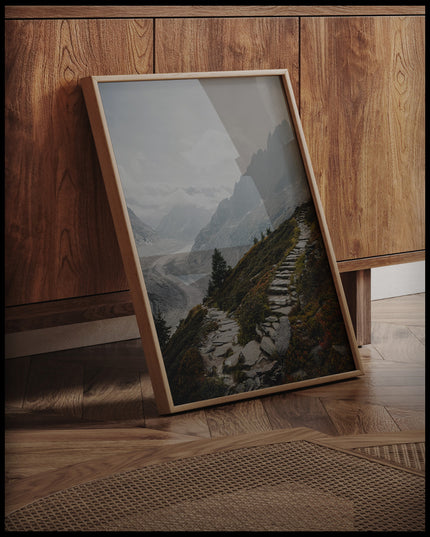 Ein gerahmtes Poster von einem Wanderweg aus steinernen Treppen inmitten einer Berglandschaft, an einer Holzkommode angelehnt und stehend auf dem Holzboden