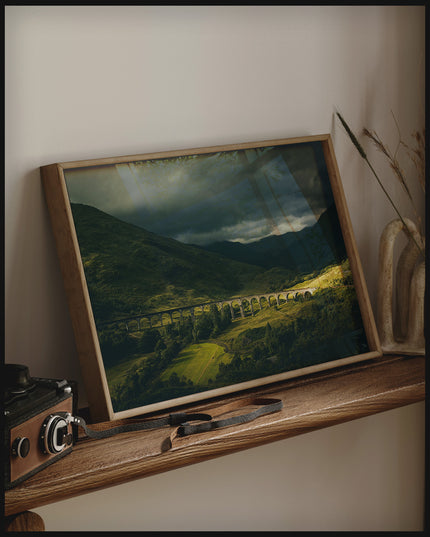 Ein gerahmtes Poster von dem Glenfinnan-Viadukt bei bewölktem Himmel, auf einem Holzregal an die Wand angelehnt