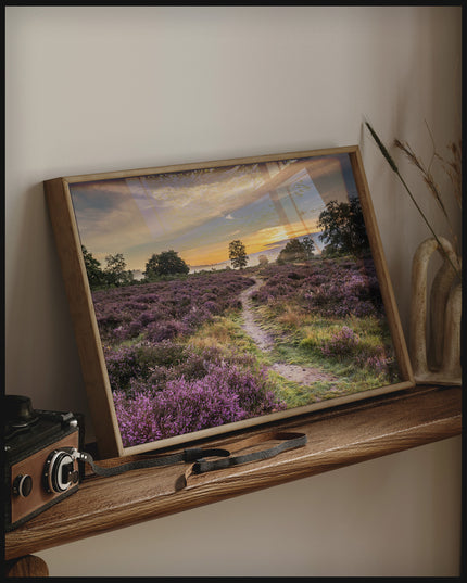 Ein gerahmtes Poster von einer Heidefläche mit lila Blumen im Sonnenuntergang, auf einem Holzregal an die Wand angelehnt