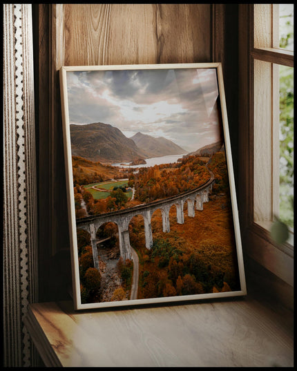 Ein gerahmtes Poster von dem Glenfinnan-Viadukt in Schottland, umgeben von herbstlicher Landschaft und Bergen im Hintergrund, vor einem Fenster an die Wand angelehnt