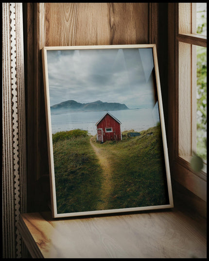 Ein gerahmtes Poster von einem kleinen roten Haus am Seeufer mit Bergen im Hintergrund, vor einem Fenster an die Wand angelehnt