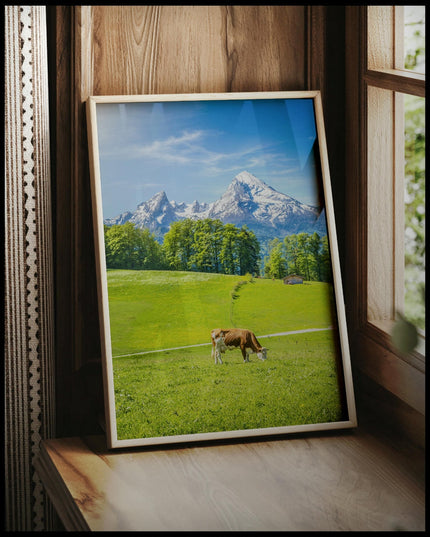 Ein gerahmtes Poster von einer Kuh auf einer grünen Wiese vor einer Alpenlandschaft, vor einem Fenster an die Wand angelehnt