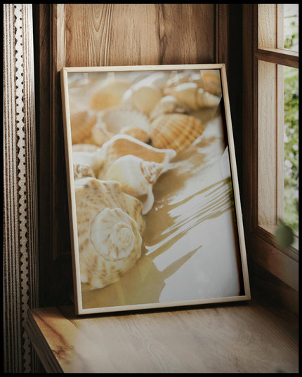 Ein gerahmtes Poster von Muscheln, die am Rand eines sandigen Strandes liegen, teilweise vom Wasser umspült, vor einem Fenster an die Wand angelehnt