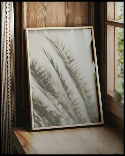 Ein gerahmtes Poster vom Schatten mehrerer Palmenblätter auf einer beigen Wand, vor einem Fenster an die Wand angelehnt