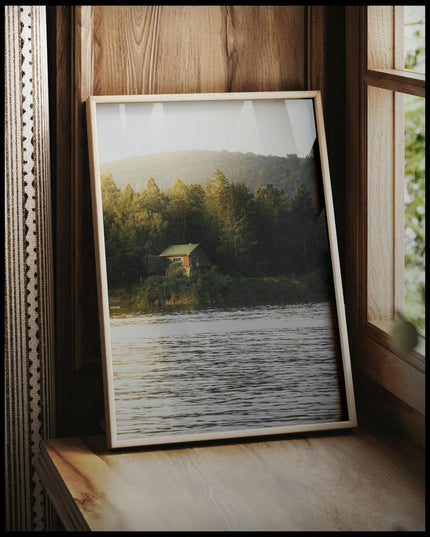 Ein gerahmtes Poster von einem Haus am See das zwischen einigen Bäumen hervorschaut, vor einem Fenster an die Wand angelehnt