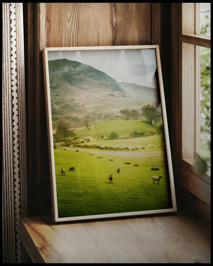 Ein gerahmtes Poster von einer grünen Weide mit Schafen vor einem Berg, vor einem Fenster an die Wand angelehnt